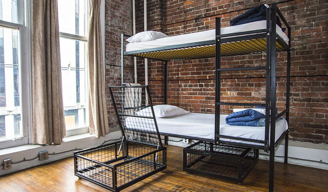 Dorm room with industrial-style metal bunk beds, tall windows, and exposed brick walls at Cambie Hostel Gastown, Vancouver